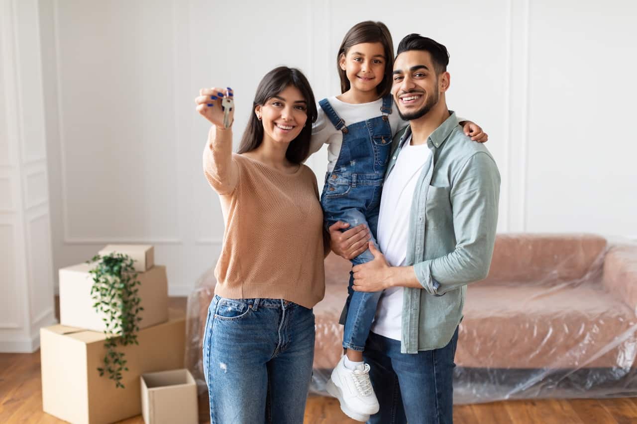 House Ownership. Portrait Of Happy Middle Eastern Family Showing Key, Standing In Living Room. Cheerful Dad Holding Daughter On Hands, People Moving In New Apartment, Posing, Blurred Background